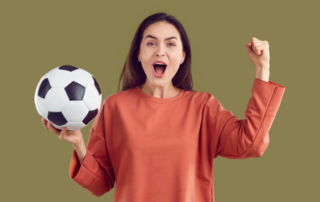 Funny, cheerful and crazy female cheerleader supporting her favorite team with soccer ball in hand. Woman shouting loudly and waving her fist while watching soccer match on khaki background. Banner.の写真素材