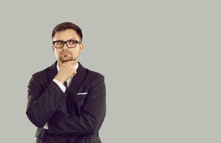 Serious pensive businessman looks at content on copy space which arouses suspicion and doubt. Young man in suit and glasses holding his chin looks to side on gray background. Isolated. Web banner.の写真素材