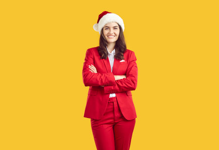 Portrait of happy business agent or event manager in red suit and Christmas hat. Happy beautiful young woman in stylish red suit and Santa cap standing with arms folded on yellow studio backgroundの写真素材