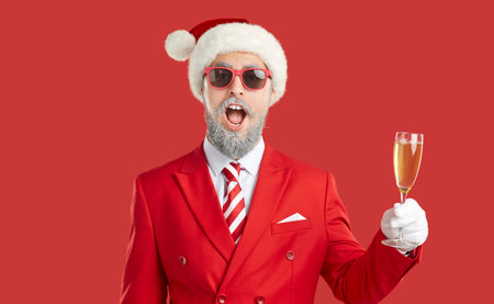 Funny man drinking champagne on Christmas day. Funny surprised bearded man in red suit, Santa hat and sunglasses standing on red studio background, raising his glass and saying toastの写真素材