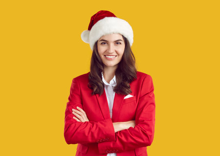Portrait of stylish beautiful woman in red santa hat on orange background in studio. Caucasian brunette woman in red elegant suit smiling looking at camera posing with folded arms. New Years banner.の写真素材