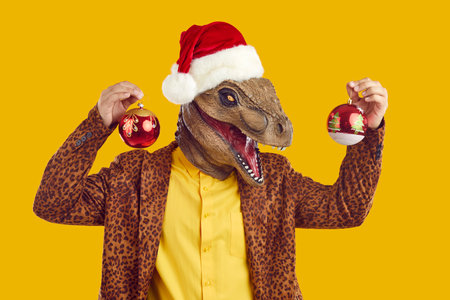 Funny joyful excited dinosaur guy with Christmas baubles. Happy man wearing dino monster mask, Santa Claus cap and funky leopard jacket holding Xmas tree balls standing isolated on yellow backgroundの写真素材