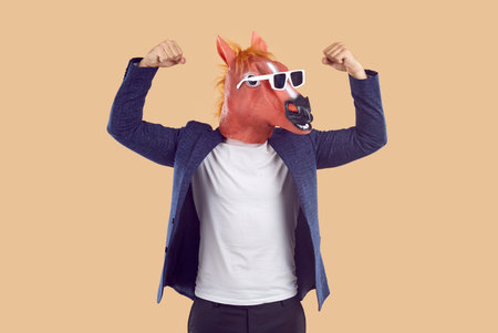 Eccentric man in funny weird costume showing his strong arms. Male model wearing business suit, bizarre fancy dress masquerade horse face mask and sunglasses pretends to be strong and flexes his armsの写真素材