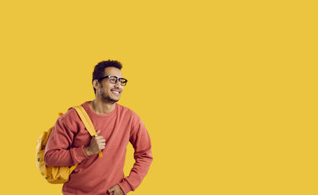 Happy cheerful college or university student with backpack isolated on copy space background. Handsome young black man in sweatshirt and glasses smiling and looking away at yellow studio backgroundの写真素材