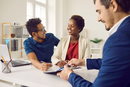 Couple of real estate buyers are ready to enter into agreement to buy apartment or house with real estate agency. Agent sitting at table shows place to sign document to happy African American family.の写真素材