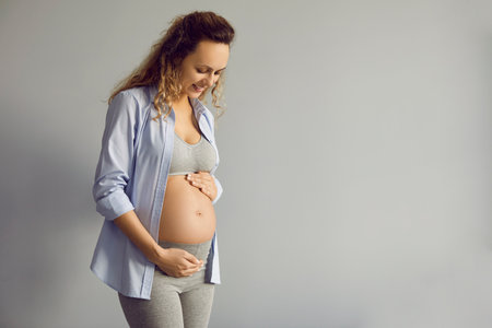 Baby expectation. Portrait of happy young woman standing on gray background and touching her hands...