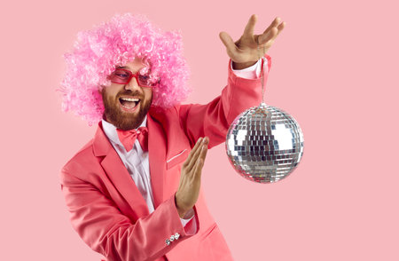 Crazy cheerful and funny man spins little shiny disco ball isolated on pink background. Young Caucasian man in pink wig, glasses and coral suit is having fun and enjoying party. Banner.の写真素材