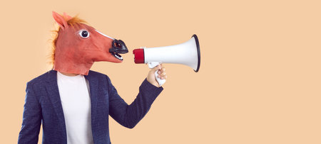 Man in trendy rubber horse mask makes announcement or advertises using megaphone. Creative concept for advertising. Man with animal head speaks into loudspeaker near copy space on beige background.の写真素材