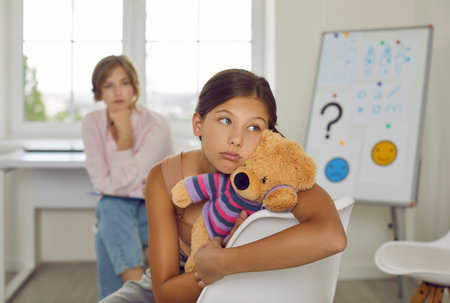 Troubled and indifferent preteen girl ignores female psychologist during therapy session. Portrait of Caucasian girl sitting on chair with her toy and turning away not wanting to talk to psychologist.の写真素材
