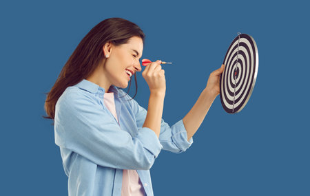 Happy beautiful confident young brunette woman in casual shirt squints eye and aims dart arrow at...