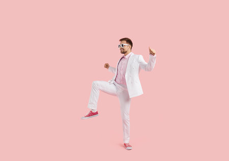 Overjoyed young man in suit isolated on pink studio background dancing feeling joyful and optimistic. Funny guy in costume and sunglasses have fun make dancer moves.の写真素材