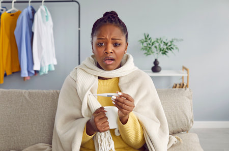 Young African American woman having cold and high fever. Unhealthy girl wrapped in plaid checking body temperature by digital thermometer. Attractive girl caught cold looking at cameraの写真素材