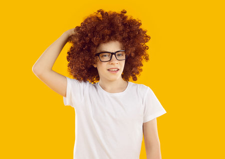 Portrait of funny puzzled child. School boy in curly brown wig and eyeglasses standing isolated on yellow background, thinking, scratching head and looking at camera with confused, unsure expressionの写真素材