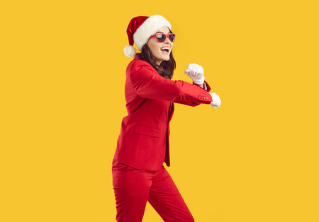Happy cheerful carefree positive confident Santa woman in funky red suit, Christmas cap, white gloves and sunglasses dancing isolated on yellow color background. Christmas fun, New Year party conceptの写真素材