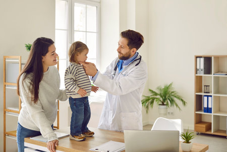 Mother and baby toddler visiting pediatrician in medical clinic. Male doctor in white coat with stethoscope examining little patient, who standing on table. Baby being checked by doctorの写真素材