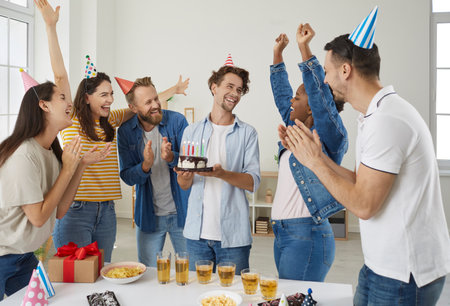 Be happy, birthday boy. Group of cheerful, excited friends having fun at a birthday party. Happy man holding a cake with candles while his diverse mixed race friends are laughing and cheeringの写真素材