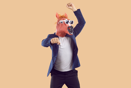 Funny unrecognizable man in rubber horse mask is fooling around dancing in gangnam style. Cheerful crazy businessman in jacket, t-shirt and pants having fun during costume party on beige background.の写真素材