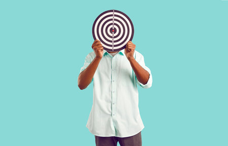 Anonymous man in casual clothes hides face behind dart board symbolizing selection of target...