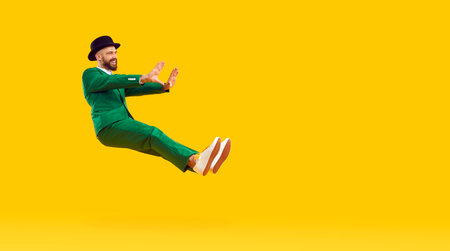 Happy funny young man wearing a green suit and a black hat floating and flying in the air isolated on a bright yellow color copy space background. Advertising, fashion, St Patricks Day conceptの写真素材