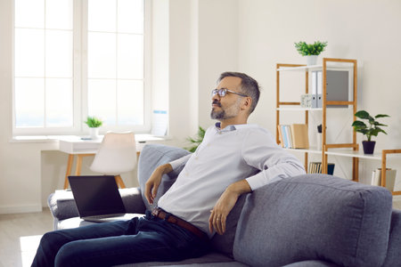Businessman relaxing on the sofa after work. Mature business man in a white shirt and glasses sitting on the couch with his laptop, relaxing and enjoying free timeの写真素材