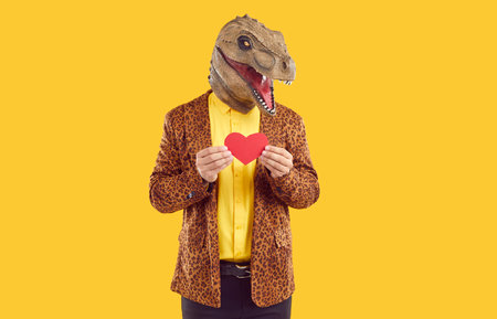 Funny Valentines Day. Dinosaur man holding red heart over isolated studio background. Portrait of funny man wearing t-rex dinosaur latex head mask showing red paper heart expressing love.の写真素材