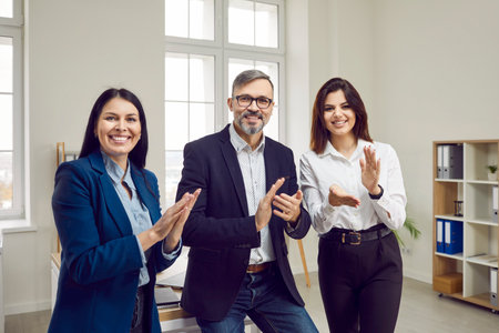 Brutal, gray-haired, happy man and two beautiful, smiling young businesswomen applaud looking at the camera. Business meeting and applause, positive atmosphere in the office.の写真素材