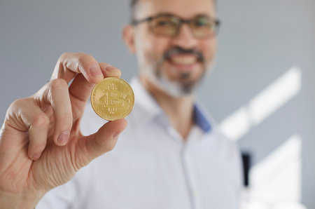 Close up of a golden bitcoin in a mans hand. Happy mature businessman holding a gold coin with a bitcoin symbol, closeup shot, soft focus. Business, crypto currency concept backgroundの写真素材