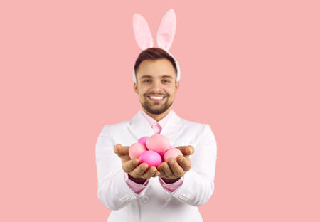 Smiling man wearing bunny ears holding Easter eggs isolated on pastel pink background. Close up of...