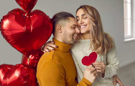 Young couple in love in romantic atmosphere is sitting among heart-shaped balloons in the living room of the house. Smiling young man gives his heart to his attractive girlfriend for Valentines day.の写真素材