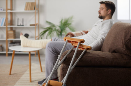 Crutchers close up on the background of a handicapped young man sitting at home on sofa in rehabilitation with his crutches. Brunet guy with injured leg with walking stick. Selective focus.の写真素材