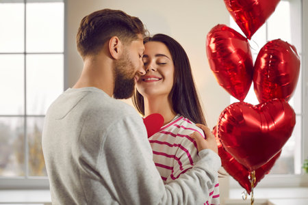 Happy man and woman celebrate Valentines Day and make romantic gifts to each other. Indoor shot of a young couple in love hugging, with red heart shaped balloons in the backgroundの写真素材
