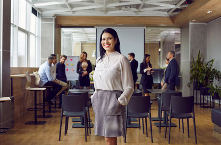 Portrait of friendly and successful business woman standing against background of colleagues in office. Millennial female leader or coach stands against background of people talking to each other.の写真素材