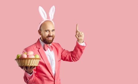 Funny bald man with ginger beard, wearing suit, bow tie and cute bunny ears standing on pink background, holding bowl of Easter eggs, smiling, pointing first finger up, suggesting fun interesting ideaの写真素材