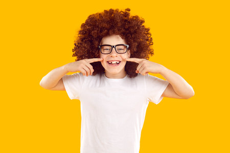 Funny happy cheerful positive school child boy in white T shirt and curly wig smiles and points with...