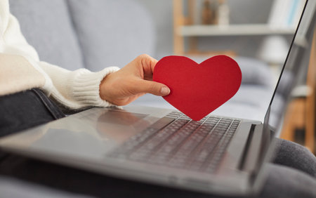 Young woman is looking for love online and using a dating website. Cropped shot of an unrecognisable lady with a laptop computer on her lap holding a red paper heart. Online dating conceptの写真素材