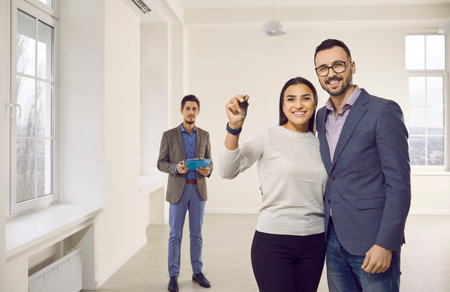 Young family couple buying or renting new house or apartment. Portrait of happy man and woman showing key while standing together in white living room at home, with real estate agent in backgroundの写真素材