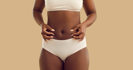 Black woman in underwear pinches her belly standing on beige color background. Plus size girl fights body fat by eating less food on weight loss diet, doing laser lipolysis treatment, and exercisingの写真素材