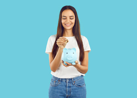 Portrait of happy woman who puts coin in piggy bank, saving money for purchase she dreams of. Young woman in casual clothes holding piggy bank in shape of pig isolated on light blue background.の写真素材