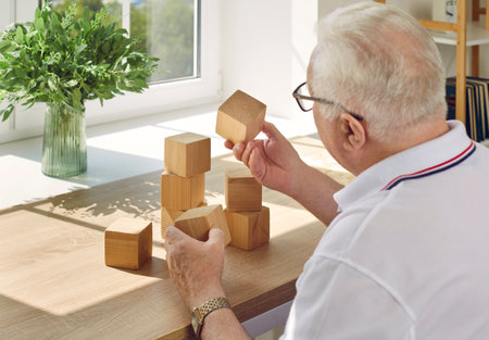 Elderly man collects wooden cubes at table opposite the window in modern rehabilitation center for the feeble-minded. Creative idea of training for dementia, feeble-minded, Alzheimers disease.の写真素材