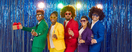 Group of cheerful friends with glasses of champagne at themed New Years disco party. Panoramic view of people in colorful clothes, retro wigs and various glasses against shiny foil curtain backgroundの写真素材