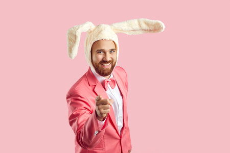 Funny young guy in Easter bunny costume points his finger at camera. Happy confident bearded man wearing plush toy rabbit ears standing on pink studio background, smiling and pointing at cameraの写真素材