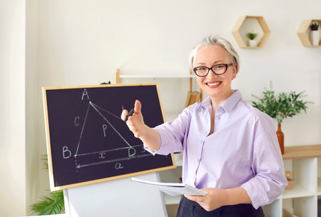 Cheerful gray-haired Caucasian woman teacher or private tutor stands at blackboard with geometric drawings for teaching schoolchildren or students exact sciences standing in classroomの写真素材