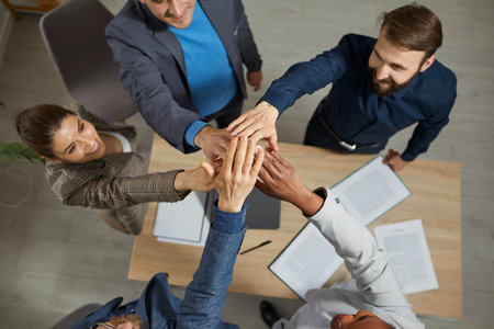 Happy business team joining hands after successful work meeting. Cheerful joyful positive multiracial people pile up hands while standing around desk in office workplace. From above. Teamwork conceptの写真素材