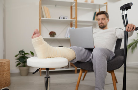 Man with broken leg sitting in armchair using laptop computer. Handsome man with plastered limb and...