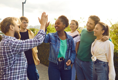 Friendly gesture. Group of happy young people give each other high five during meeting on city street. Cheerful African American woman greets her Caucasian male friend surrounded by their best friendsの写真素材