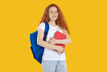 Portrait of cheerful teenage girl with notebooks and school backpack isolated on orange background....