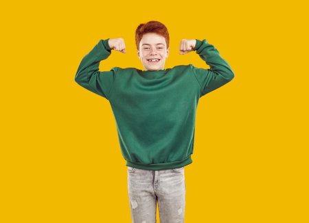 Red-haired, happy, smiling, confident teenager, dressed in green sweater and gray jeans, demonstrates his strength and biceps on both arms. Isolated studio photo on bright yellow background.の写真素材