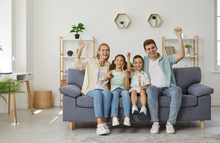 Happy family with two children on couch watching sport game on TV and and they happy about winning of their team. They laugh, shout and raise their hands up. Family lifestyle, spending time together.の写真素材