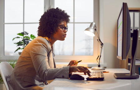 Professional accountant working in office. Side, profile view of young African American woman sitting at desk, looking at screen of desktop computer, using digital business sheets, working with dataの写真素材