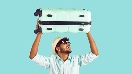 Happy Indian man having fun lifting travel suitcase above his head on light blue background. Joyful bearded man in shirt, sunglasses and summer hat goes on journey. Concept of travel and vacations.の写真素材